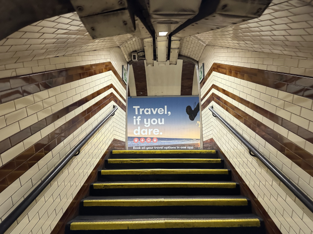 Eine Treppe in einer U-Bahn-Station mit einem Schild, das oben 'Reise, wenn du wagst' trägt, flankiert von Geländern auf beiden Seiten und sichtbaren Stationswänden im Hintergrund.