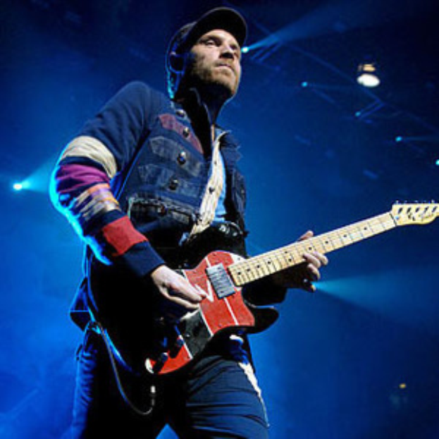 Ein Mann mit einer Mütze spielt Gitarre auf der Bühne eines Konzerts, beleuchtet von hellen Scheinwerfern, mit einem freudigen und aufgeregten Gesichtsausdruck.