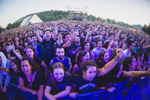 Menschenmenge auf einem Musikfestival mit erhobenen Händen vor einem Geländer, Bäume und Strukturen im Hintergrund unter einem klaren blauen Himmel.