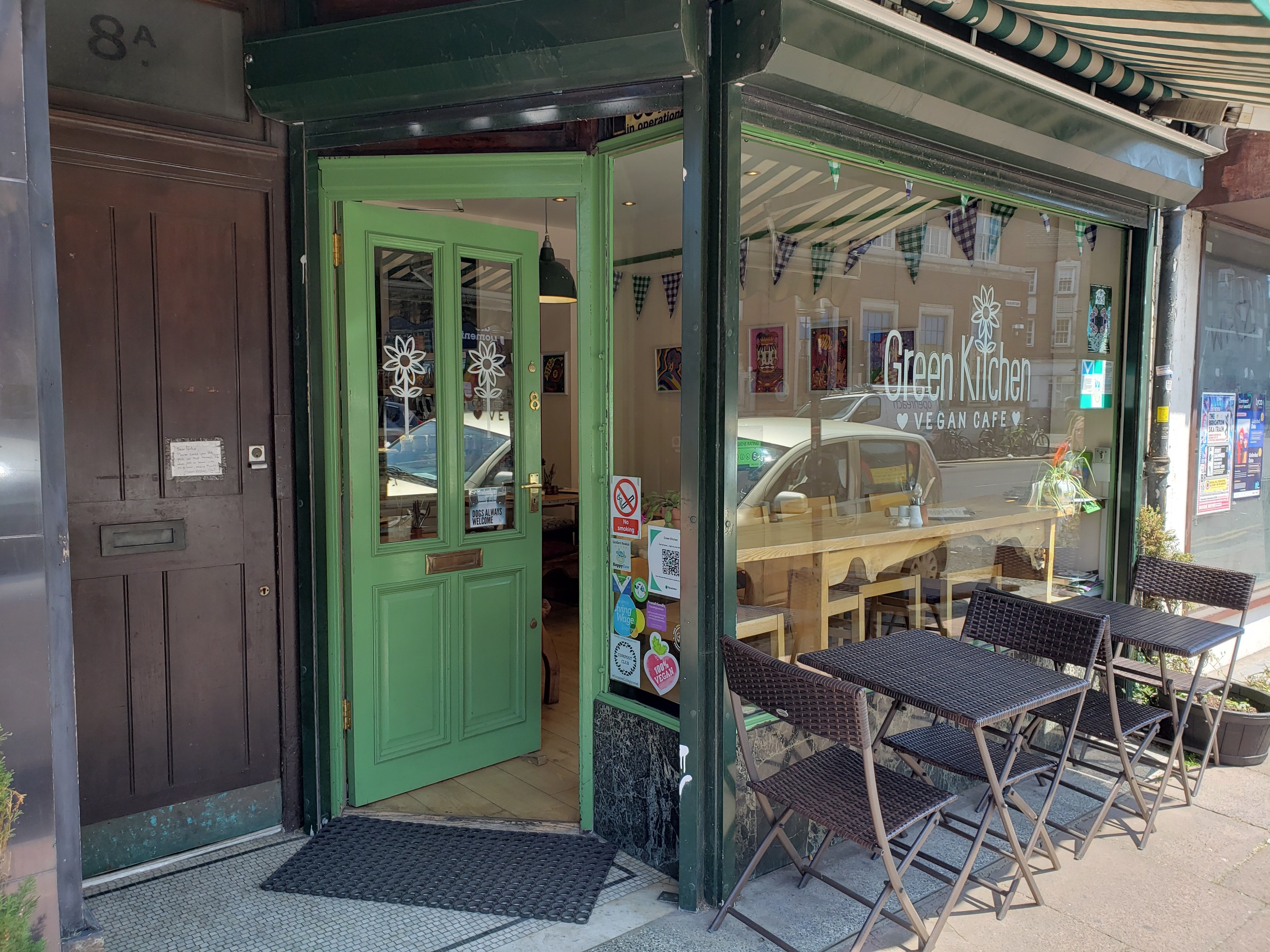 Außensitzbereich des Green Kitchen Vegan Cafe mit Tischen, Stühlen, Pflanzen, einer Matte und einer Glastür mit Postern.