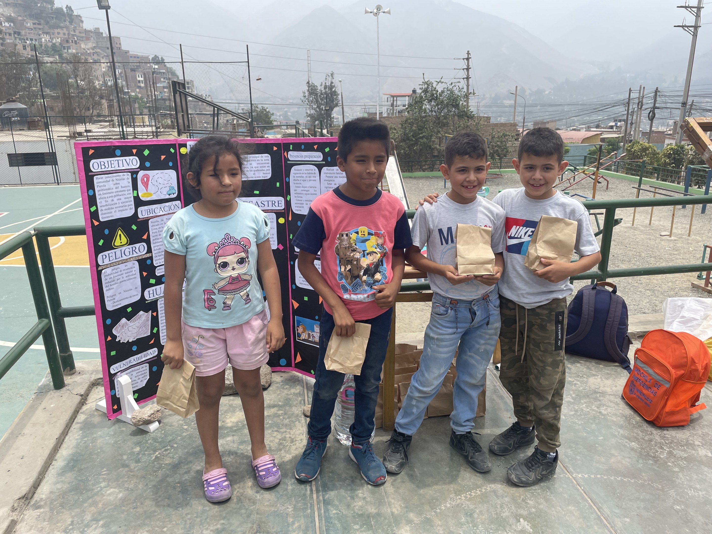 Gruppe von Kindern auf einem Basketballfeld, die Papierbeutel halten, mit Beuteln und Gegenständen rechts und einer Tafel mit Text links, vor einem Hintergrund aus Zaun, Pfosten, Straßenlaternen, Bäumen, Gebäuden, Hügeln und Himmel.