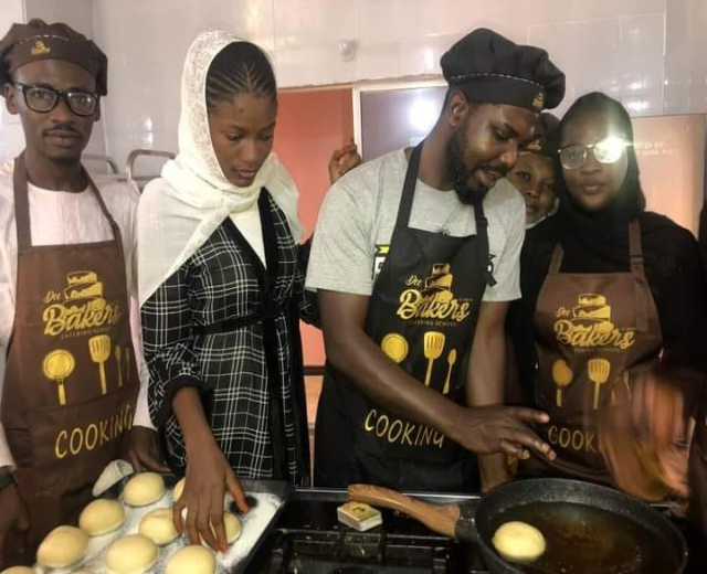 Eine Gruppe von Menschen in Schürzen und Mützen bereitet in einer Küche mit einem Topf auf dem Herd, Schränken und einer Wand im Hintergrund Essen zu.
