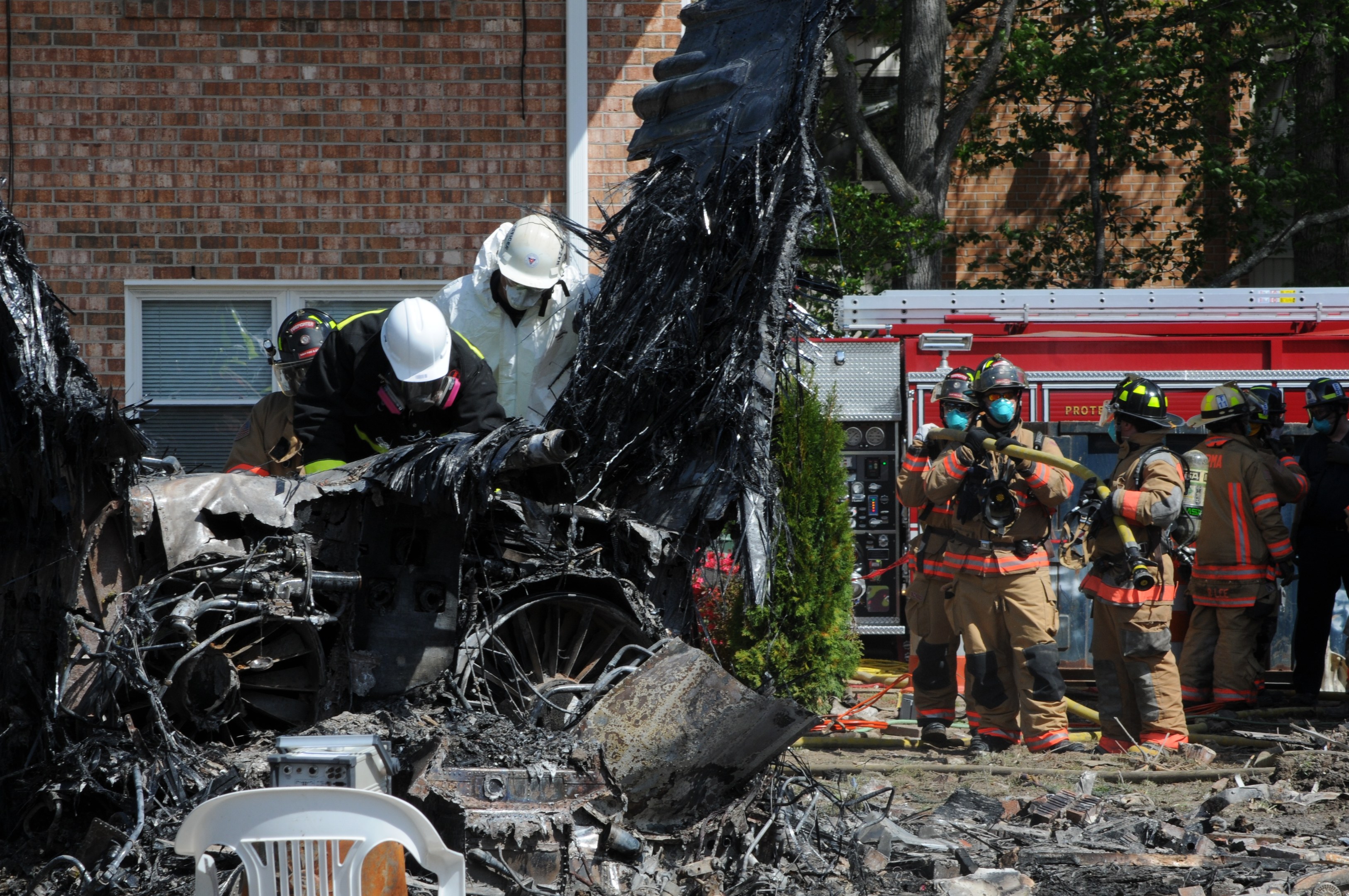 Feuerwehrleute in Schutzausrüstung löschen ein Gebäude Feuer, während verkohlte Trümmer, einschließlich eines Fahrzeugs und verstreuter Gegenstände, den Boden hinter ihnen bedecken.