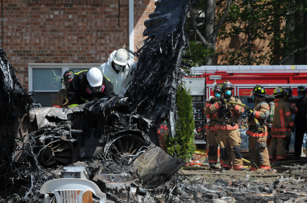 Feuerwehrleute in Schutzausrüstung löschen ein Gebäude Feuer, während verkohlte Trümmer, einschließlich eines Fahrzeugs und verstreuter Gegenstände, den Boden hinter ihnen bedecken.