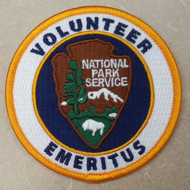 Ein rundes Abzeichen mit einem blauen Rand, weißem Hintergrund und fetter schwarzer Schrift, die 'Volunteer National Park Service Emeritus' in der Mitte eines gelben Kreises liest.