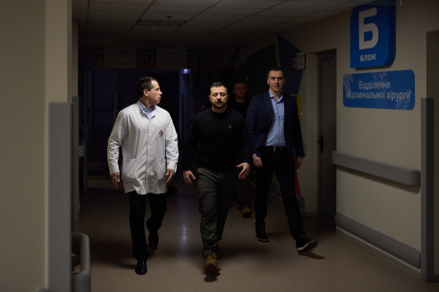 Eine Gruppe von Männern geht einen hell erleuchteten Krankenhausflur entlang, mit einer Tür auf der rechten Seite und Texttafeln auf der linken Seite, die besorgt wirken.