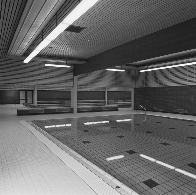 Innenpool mit klarem Wasser, gefliestem Boden und Wänden, Deckenbeleuchtung und einigen umliegenden Gegenständen in einem großen, ruhigen Gebäude.