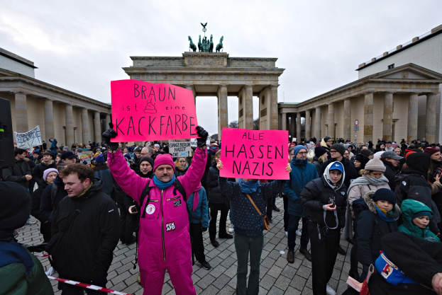 Gruppe von Menschen protestiert vor dem Brandenburger Tor in Berlin, hält Schilder gegen die Anti-Nazi-Bewegung der deutschen Regierung, mit Gebäuden, Säulen und Statuen im Hintergrund unter einem klaren blauen Himmel.
