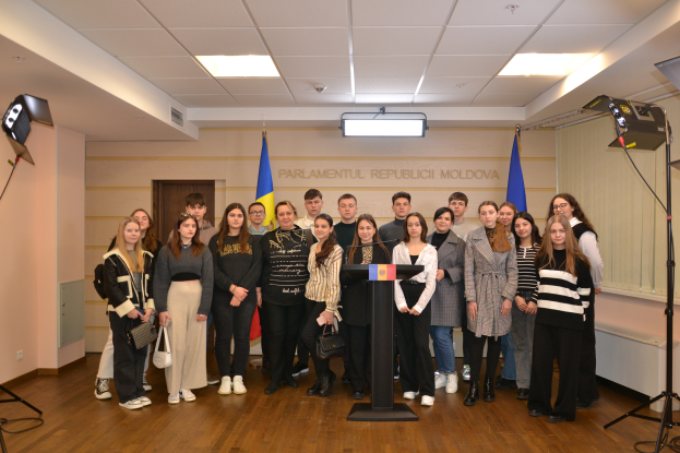 Eine Gruppe von Menschen steht vor einem Podium mit Mikrofon, umgeben von Fahnen, mit einer Wand, die "Moldova Youth Congress 2018" anzeigt, einer Tür, einem Dach mit Deckenleuchten und Ständern mit Leuchten.