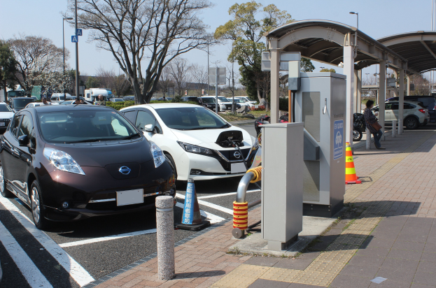 Ladestation für Elektroautos in Japan mit Autos, Verkehrskegeln, einer Person, einem Schuppen, Mästen, Lichtern, Schildern, Bäumen, Pflanzen und einem Himmel im Hintergrund.