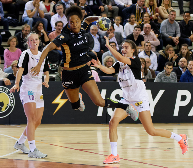 Eine Gruppe von Frauen beim Handballspielen auf einem Platz mit einem Ball in der Mitte, Zuschauern im Hintergrund und einer Fahne mit Text und Logos.
